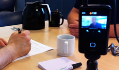 En bruger ses gennem et videokameras display, mens en researcher tager noter.